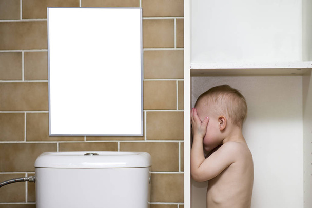 Blank poster above a toilet with a boy standing in a bathroom cabinet covering his eyes with his hands with space for text or picture or product placement above and next to him. With clipping paths - Photo, Image