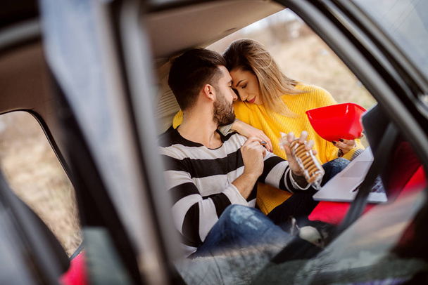 Porträt eines glücklichen jungen Paares, das sich einen Film auf dem Laptop anschaut und Popcorn isst, im Auto picknickt - Foto, Bild