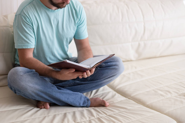 Mann liest altes gebundenes Notizbuch in den Händen haltend auf der weißen Couch. Inländischer Lebensstil. Bildungskonzept. - Foto, Bild
