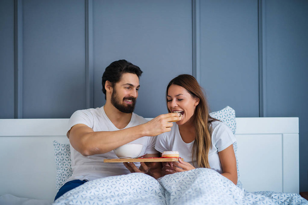 houkutteleva nuori onnellinen mies ruokinta tyttöystävä sängyssä kotona
 - Valokuva, kuva