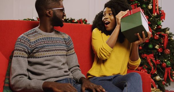 Jeune couple afro-américain amoureux assis sur le canapé rouge dans la chambre décorée de Noël confortable avec un beau sapin de Noël derrière eux. bel homme remis cadeau de Noël à sa petite amie et
 - Photo, image