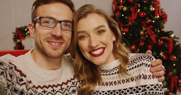 Joven pareja atractiva que tiene videochat en la bonita sala de estar con decoraciones de Navidad y un árbol de Navidad. En el interior. De cerca. Retrato. POV
 - Foto, imagen