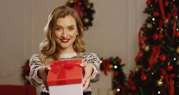 Portrait shot of young blonde beautiful woman smiling and handing a present with a red ribbon to the camera. The Christmas tree with lights behind. Inside. Close up - Foto, immagini