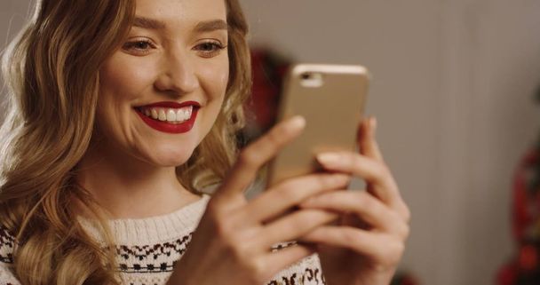 Close up of young smiled and happy blonde woman taping and chatting on her mobile phone and making surprised face. Christmas tree on the background. Portrait shot. - Photo, image
