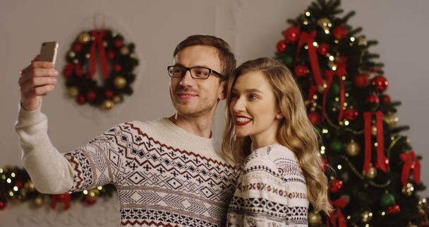 Young happy attractive couple in love taking nice photos on a smart phone and posing in the Christmas atmosphere of living room. Selfies on the Christmas tree background. Indoor - Фото, зображення