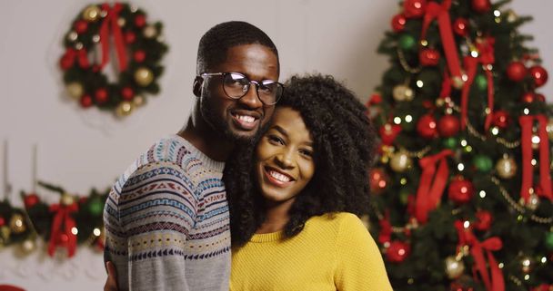 Portret van jonge en gelukkig Afrikaanse Amerikaanse echtpaar kijken naar elkaar en glimlachend in de camera op een kerstboom met lampjes achtergrond. Binnenshuis. - Foto, afbeelding