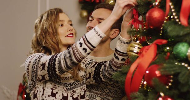 Young attractive happy couple decorating the Christmas tree with red ribbons and golden baubles. Inside the living room. Close up. Portrait - Фото, изображение