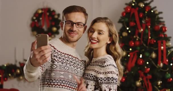 Portrait of young happy couple in love making smiling selfies on a smart phone and showing victory signs in the nice decorated living room. Christmas tree background. - Filmati, video