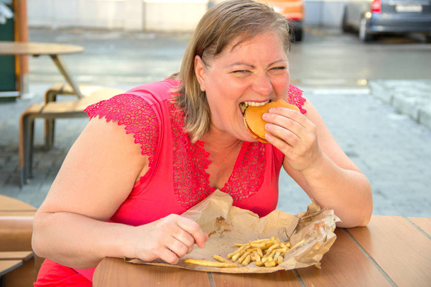 Paksu nainen syö pikaruokaa hampurilainen ja ranskalaisia perunoita kahvilassa
 - Valokuva, kuva