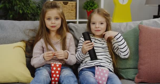 Two cute little girls laughing while sitting on the couch with popcorns and watching TV with a remote control in hands in the cozy living room. Indoor - Imágenes, Vídeo