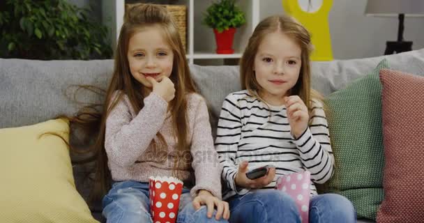 Two funny small girls with long hair eating popcorn while sitting on the sofa and watching TV with a remote control in hands in the cozy nice room. Indoors. Portrait shot - Filmagem, Vídeo