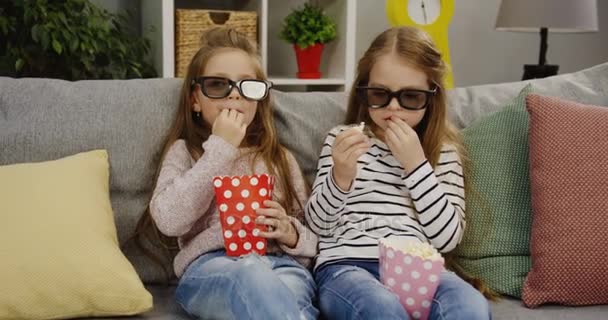 Two funny pretty little girls with long hair and wearing glasses sitting on the sofa with pillows and eating popcorn. Cute friends. Comfortable living room. Indoor - Séquence, vidéo