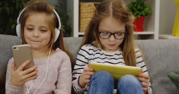 Two cute small girls sitting on the couch in the living room. One girl in the big headphones listening to the music on the smartphone and another using a tablet device. At home. Indoors - Imágenes, Vídeo