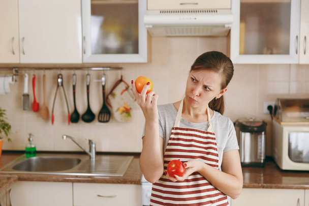 Zmatená dívka v zástěře se rozhodne vybrat červené nebo žluté rajče v kuchyni. Pojem dieta. Zdravý životní styl. Vaření doma. Příprava potravin. - Fotografie, Obrázek