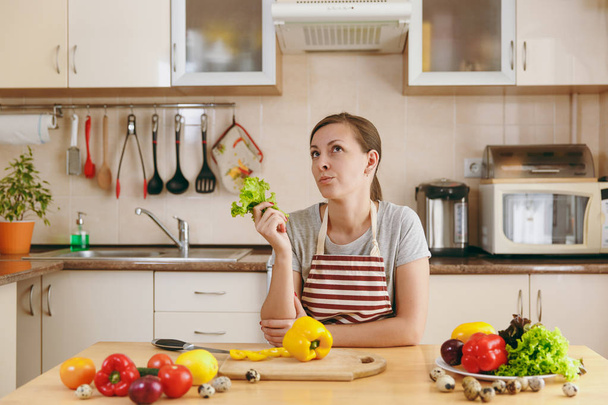 Una joven atractiva mujer pensativa en un delantal con hojas de lechuga en la cocina. Concepto de dieta. Estilo de vida saludable. Cocinar en casa. Preparar alimentos
. - Foto, Imagen