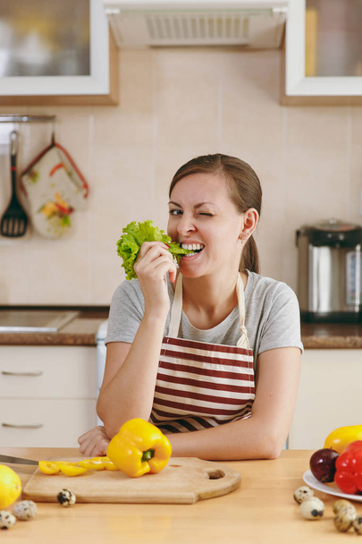 Una joven mujer guapa en un delantal con hojas de lechuga en la boca en la cocina. Concepto de dieta. Estilo de vida saludable. Cocinar en casa. Preparar alimentos
. - Foto, Imagen