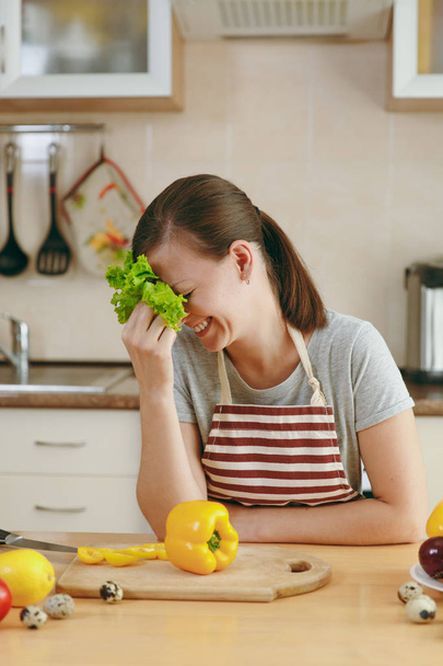 Una giovane donna attraente in grembiule con foglia di lattuga che ride in cucina. Il concetto di dieta. Uno stile di vita sano. Cucinare a casa. Preparare il cibo
. - Foto, immagini