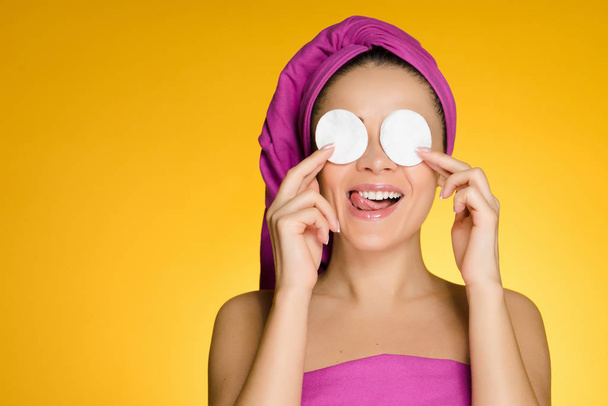 happy young girl with a pink towel on her head cleans the skin on her face with white cotton pads on a yellow background - Foto, afbeelding