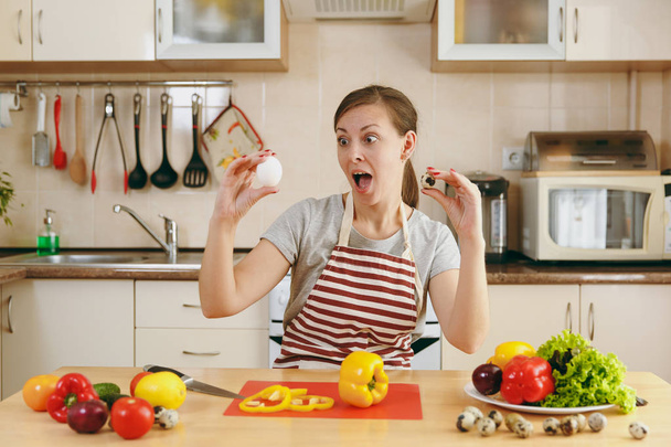 Uma jovem atraente mulher pensativa em um avental escolhe entre ovos de galinha e codorniz na cozinha. Conceito de dieta. Estilo de vida saudável. Cozinhar em casa. Preparar alimentos
. - Foto, Imagem