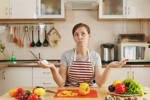 A young attractive pensive woman in an apron chooses between chicken and quail eggs in the kitchen. Dieting concept. Healthy lifestyle. Cooking at home. Prepare food. - Fotografie, Obrázek