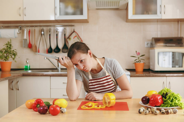 Una giovane donna stanca emaciata in un grembiule che cucina in cucina a casa. Il concetto di dieta. Uno stile di vita sano. Preparare il cibo
. - Foto, immagini