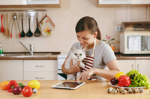 Una joven mujer bonita con gato persa blanco en la cocina con la tableta en la mesa. Ensalada de verduras. Concepto de dieta. Estilo de vida saludable. Cocinar en casa. Preparar alimentos
. - Foto, Imagen