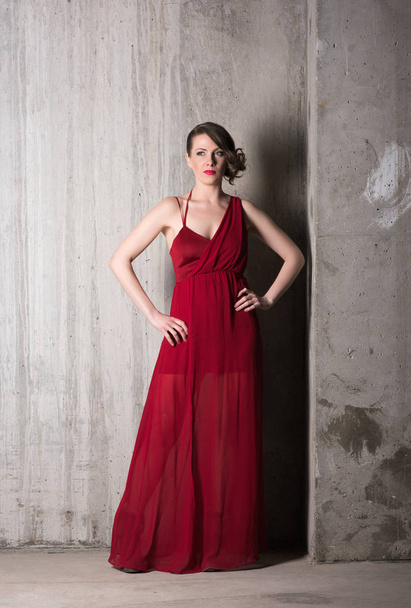 Vertical portrait of a beautiful young woman in long red dress in full growth on a background of gray concrete walls - Foto, afbeelding