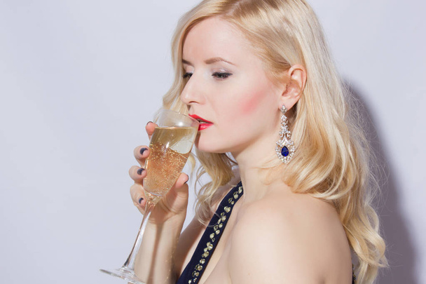 Studio portrait of a beautiful young blonde woman in a blue long evening dress. A girl is holding a glass of champagne in her hand - Foto, Bild