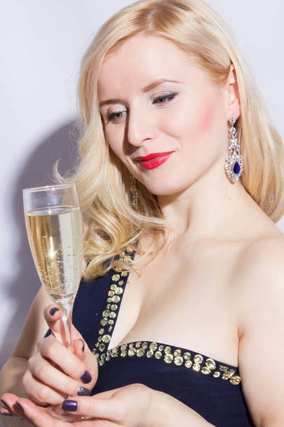 Studio portrait of a beautiful young blonde woman in a blue long evening dress. A girl is holding a glass of champagne in her hand - Fotó, kép