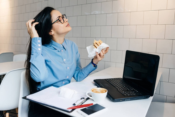 a young busy freelancer girl works in a cafe for a laptop, has a sandwich and coffee - Foto, Imagem