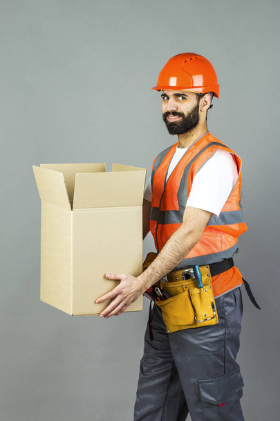 A man-builder in an orange helmet with a cardboard box in his hands. On a gray background. - Fotoğraf, Görsel