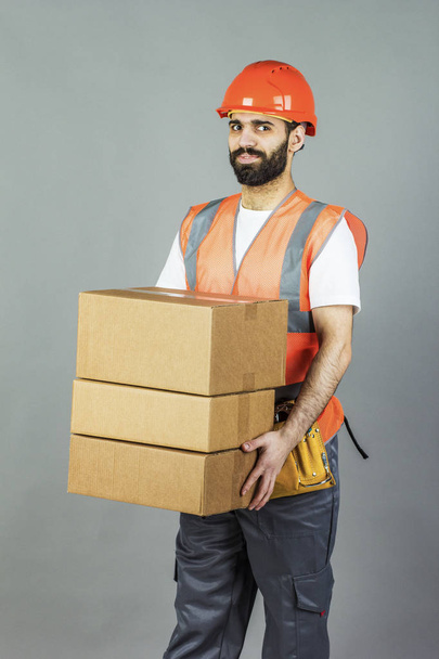 Человек-строитель в оранжевом шлеме с картонной коробкой в руках. На сером фоне
. - Фото, изображение