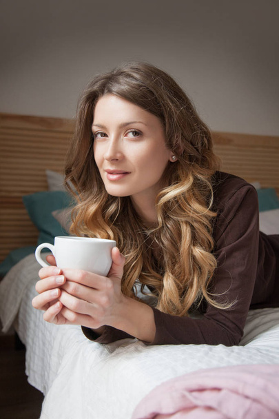 白いカップの手でベッドに横たわっている茶色のパジャマで、若い女性。朝コーヒーを飲む女性. - 写真・画像