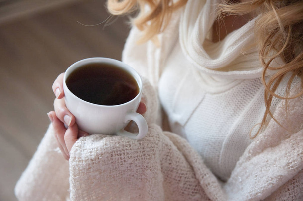 Обрізана секція молодої жінки в білому пуловері, що сидить на дерев'яній підлозі з білою чашкою чаю в руках
 - Фото, зображення