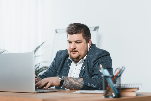 overweight businessman listening music with earphones while working on laptop in office - Photo, image