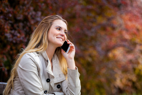 Happy woman talking smartphone sitting on bench at colorful autumn street - Zdjęcie, obraz