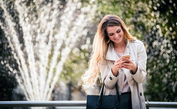 Smiling woman wearing coat walking in city and using smartphone - Photo, image