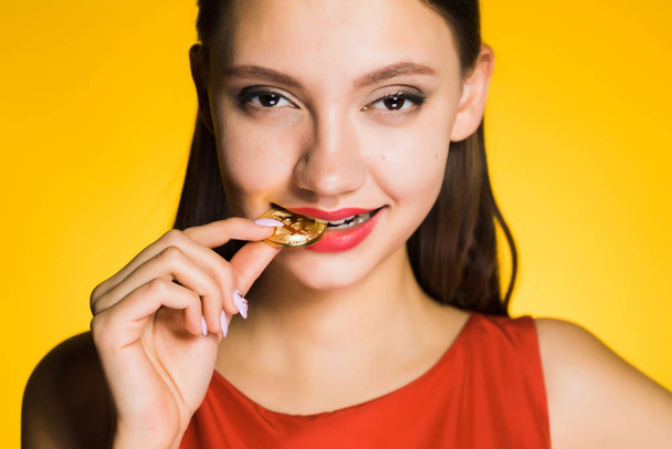 confident happy girl tries on taste golden bitcoin, on a yellow background - Фото, изображение