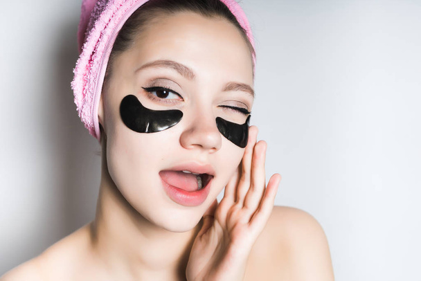 a funny young girl looks after the skin on her face, under the eyes moisturizing patches, on her head a pink towel - Foto, Bild