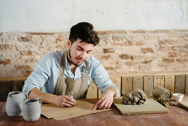 hymyilevä komea nuori meksikolainen savenvalaja piirtää paperille keramiikkaa. käsite pienyritys, käsityönä, harrastus työ
.  - Valokuva, kuva