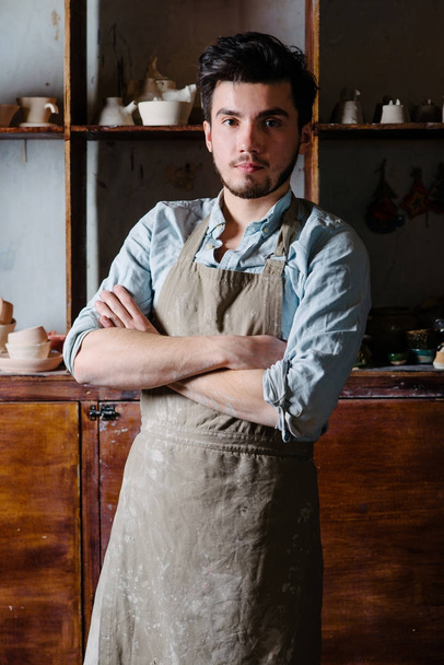 potter, tools, ceramics art concept - handsome young brunette male with a beard dressed in an apron stand with finished utensils, smiling ceramist at workplace with baked cups and jugs at background - Photo, Image