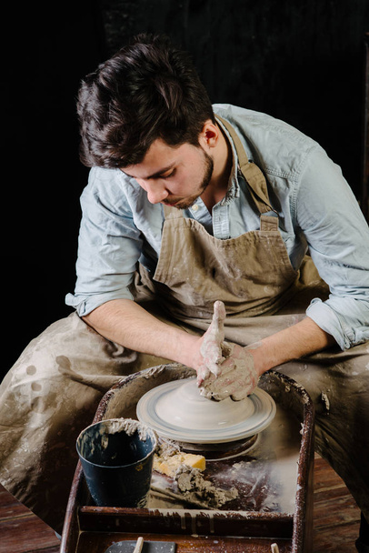 ceramica, laboratorio, concetto di arte ceramica - uomo che lavora alla ruota del vasaio con argilla cruda con le mani, una bruna maschio scolpisce un utensile vicino al tavolo di legno con strumenti, maestro in grembiule e una camicia
 - Foto, immagini