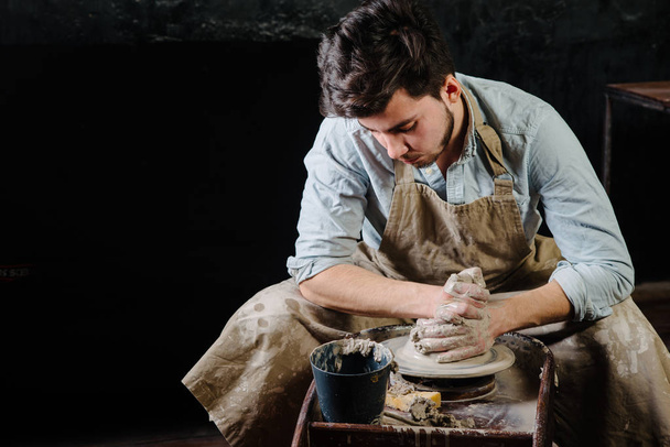 poterie, atelier, concept d'art céramique - homme travaillant sur la roue du potier avec de l'argile brute à la main, une brune mâle sculpte un ustensile près d'une table en bois avec des outils, maître en tablier et une chemise
 - Photo, image