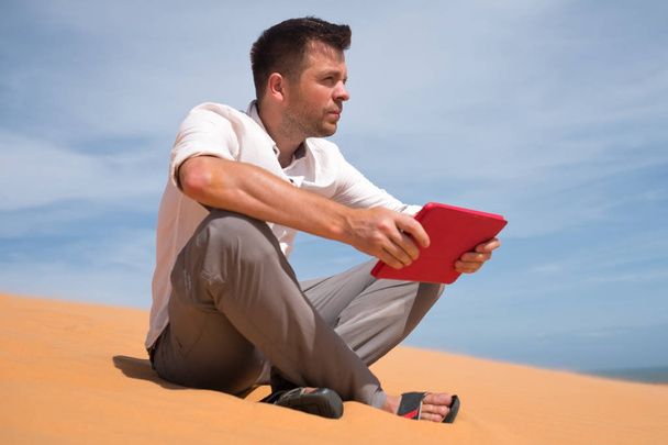 Eksyksissä aavikolla. Valkoihoinen mies istuu hiekalla tabletin kanssa. Hän on eksynyt ja etsii kartalta minne mennä.
 - Valokuva, kuva