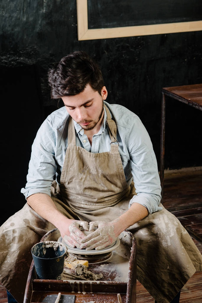 pottery, workshop, ceramics art concept - man working on potter's wheel with raw clay with hands, a male brunette sculpt a utensils near wooden table with tools, master in apron and a shirt - 写真・画像
