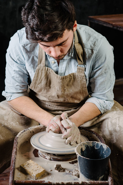 pottery, workshop, ceramics art concept - man working on potter's wheel with raw clay with hands, a male brunette sculpt a utensils near wooden table with tools, master in apron and a shirt - Photo, Image