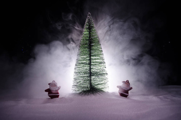 Ευτυχισμένος Βασίλη κούκλα την περίοδο των Χριστουγέννων με το δέντρο και το χιόνι. Bokeh πολύχρωμο φόντο. Santa Clause και καλά Χριστούγεννα παιχνίδι φιγούρα μοντέλο στο σκοτάδι τονισμένα - Φωτογραφία, εικόνα