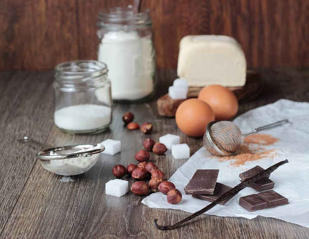 Ingredients for cooking baking (cookies or cake) on a wooden background. Flour, eggs, chocolate, hazelnut, sugar, vanilla and butter on wood table. Rustic background - Photo, image