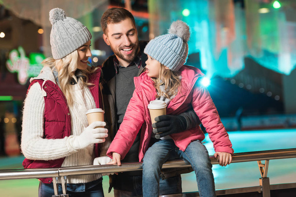 glückliche junge Familie hält Pappbecher in der Hand und lächelt einander auf der Eisbahn zu - Foto, Bild