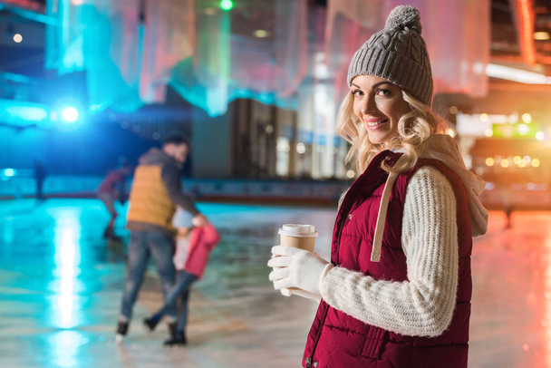 young woman holding coffee to go while her family skating behind on rink - Photo, Image
